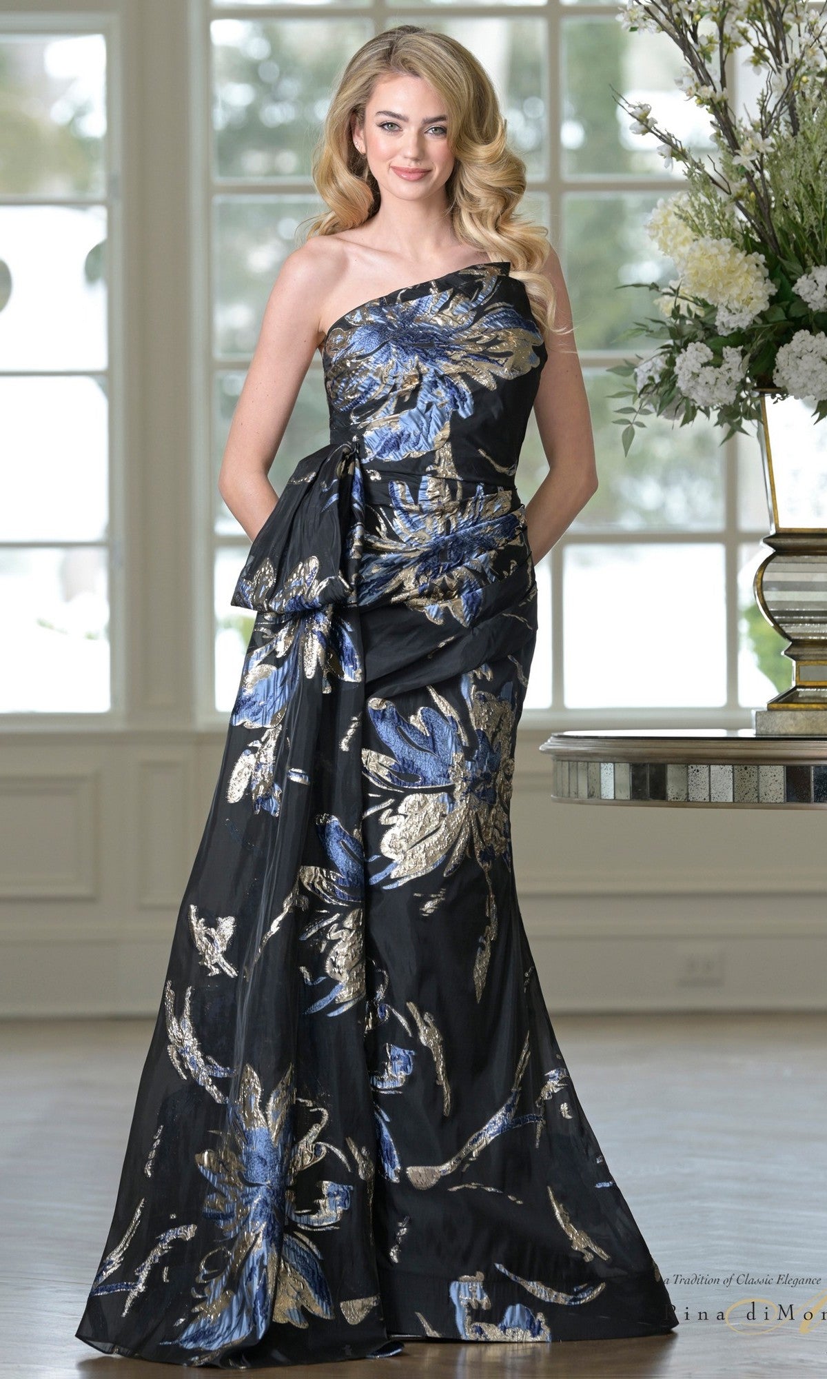 A woman smiles indoors, hands in the pockets of the Rina Di Montella RD3233 one-shoulder formal gown, featuring a black base with bold blue and gold metallic floral print.